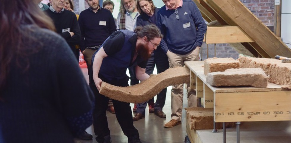 A person pushing a piece of insulation into a demo stand of a building, while a group of people look onwards