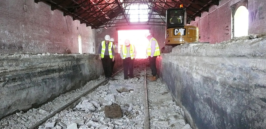 Three people standing on railway tracks inside the Engine Shed building. The tracks are flanked by two high, concrete platforms